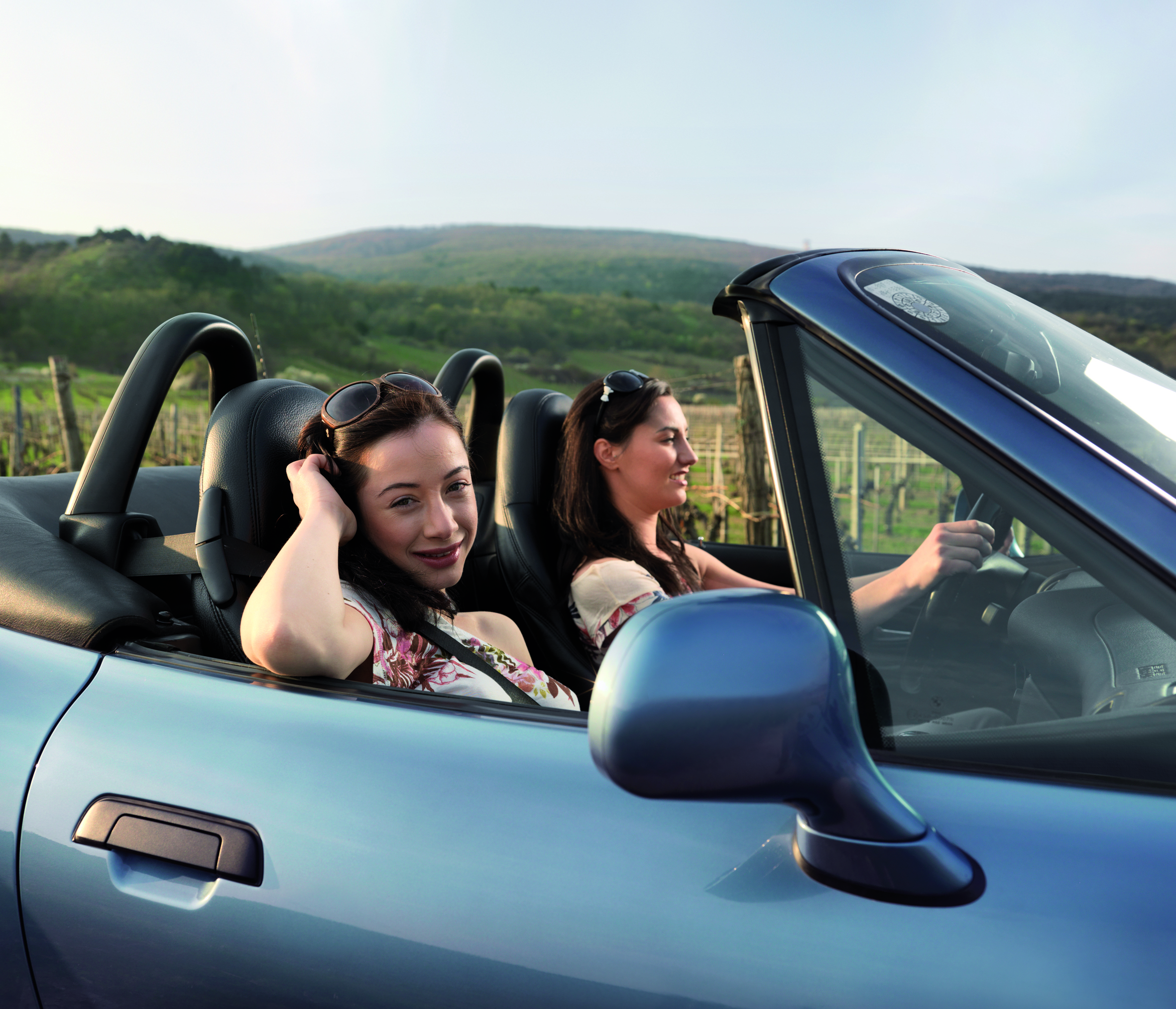 Dvije mlade djevojke u kabriolet automobilu se voze sa osmijehom, a u pozadini se vidi lijep pejzaž vinograda.